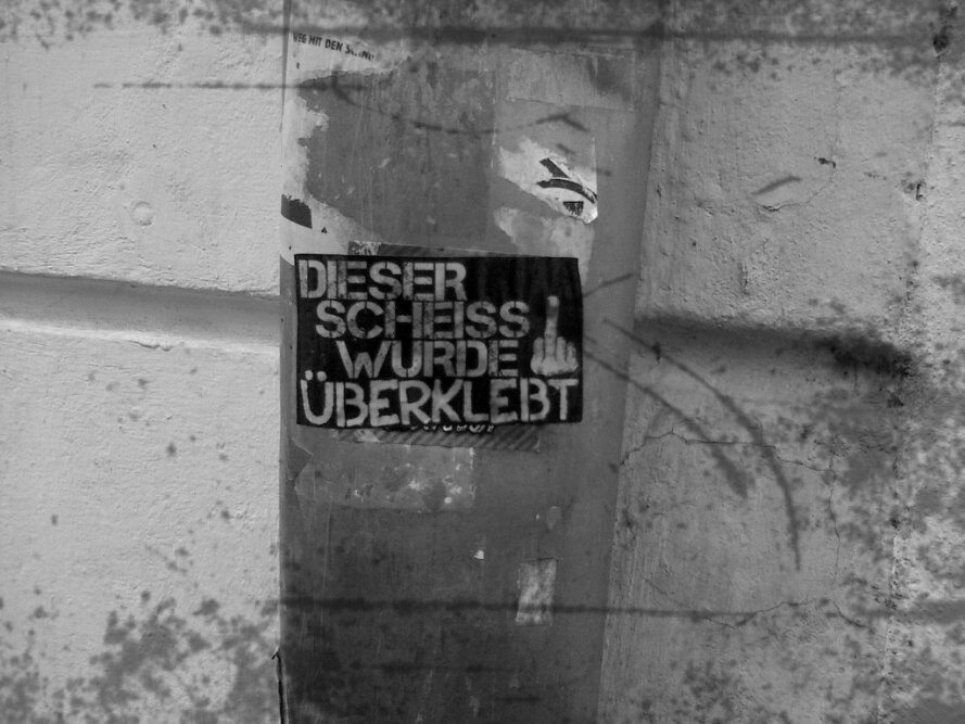 Schwarz-weißes Foto eines Stickers an einer verwitterten Steinsäule. Der schwarze Aufkleber zeigt in weißer Blockschrift den Text „DIESER SCHEISS WURDE ÜBERKLEBT" mit einem Stinkefinger-Symbol zwischen den Zeilen. Darunter und darüber sind Reste älterer, abgerissener Sticker zu erkennen.